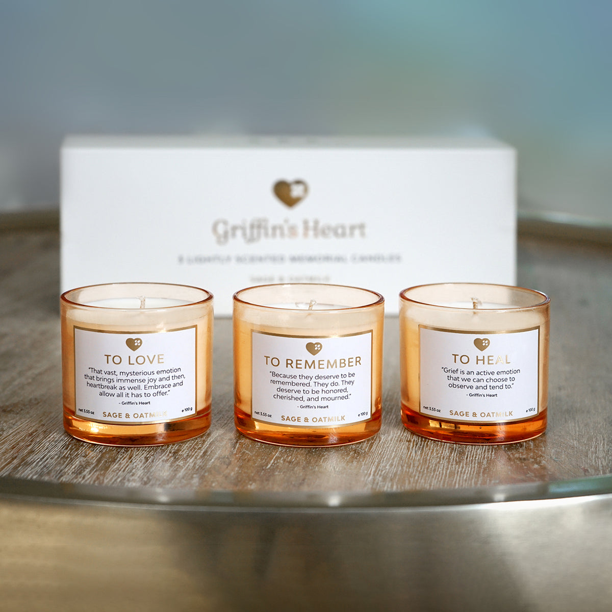 Daytime beauty shot of the Griffin's Heart Trio Candle Set on a wood and brass table, showing the elegant peach-tinted glass jars and gold detailing, SKU: GH002.