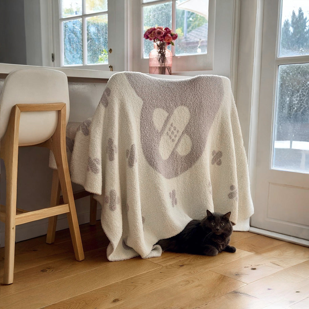 Lifestyle view of the Griffin's Heart Luxe Throw Blanket in reversible Dove Gray and White, featuring the signature band-aid heart, draped over a stool as a premium pet sympathy gift.